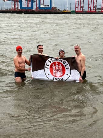 vier Personen in Badeklamotten stehen im kaltem Wasser und halten Flagge vom FC St. Pauli hoch